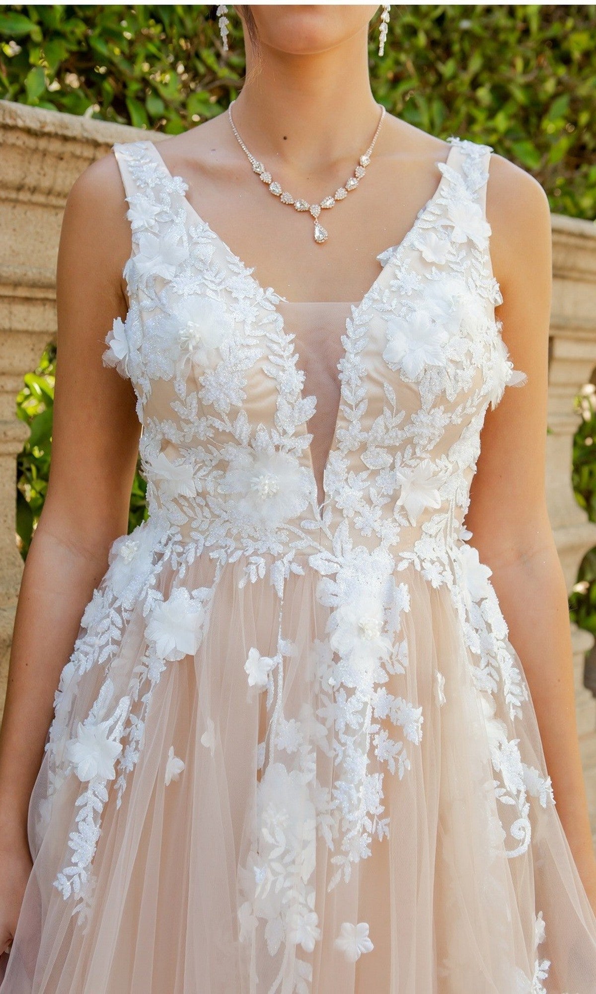 A woman wearing the Cinderella Couture 8040J long formal dress, featuring a sleeveless V-neck tulle design adorned with 3D floral lace appliques, stands outdoors in front of a stone wall and greenery.